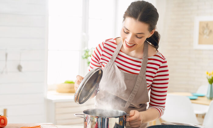 Una donna in cucina con il grembiule solleva il coperchio di una pentola. Sul piano di lavoro si vedono pomodori tagliati a fette.