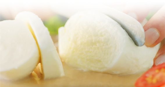 La mozzarella GOLDSTEIG viene tagliata a fette su una tavola di legno. Pomodoro e basilico sullo sfondo.